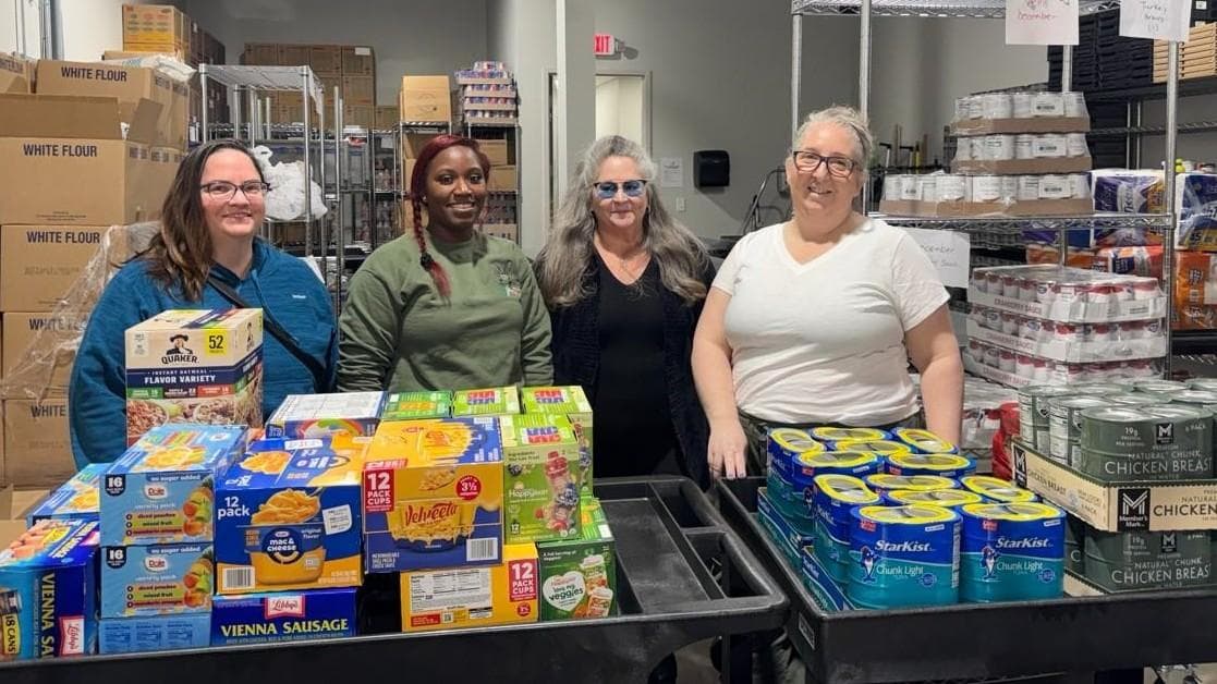 Four women stand together surrounded by boxes and shelves of shelf stable foods.
