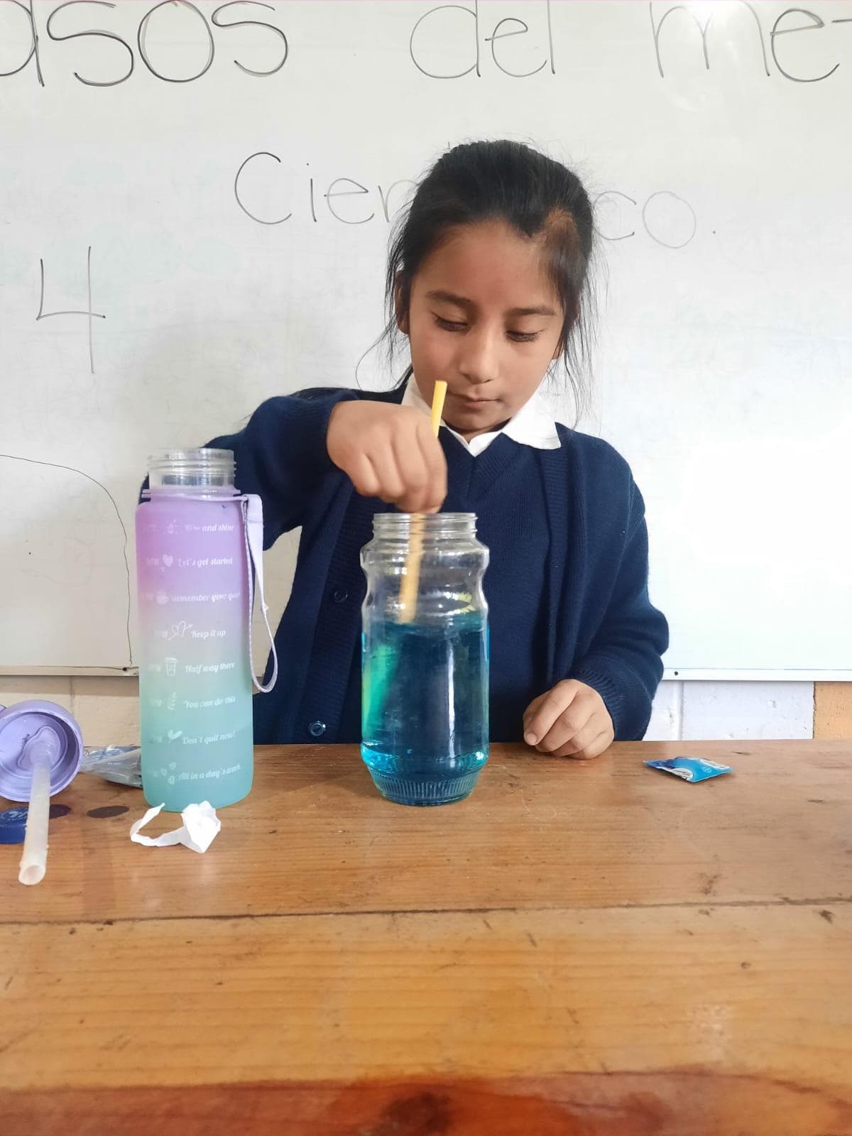 An elementary school student wearing her school uniform mixes blue colored liquid in a jar on a desk in front of a whiteboard.
